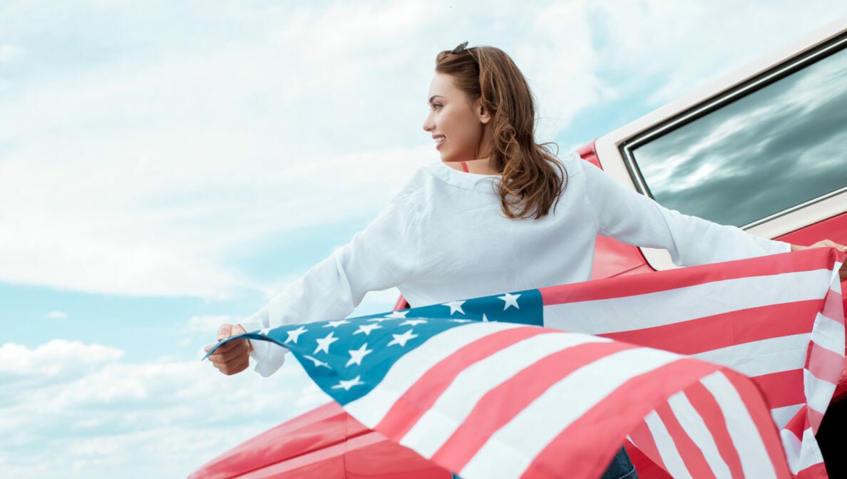 Eine weibliche Person in Rückansicht vor einem roten Auto, die eine amerikanische Flagge hinter sich wehen lässt.