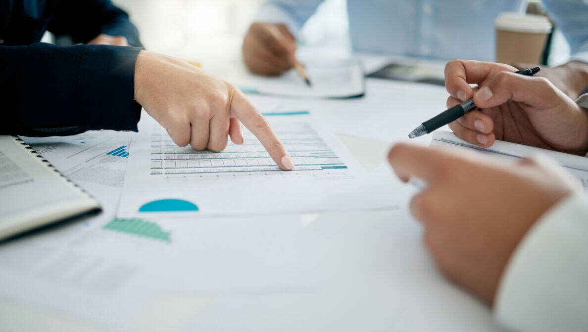 Drei Personen an einem Tisch. Eine Person zeigt auf ein Papier in der Mitte, auf dem Tabellen abgebildet sind