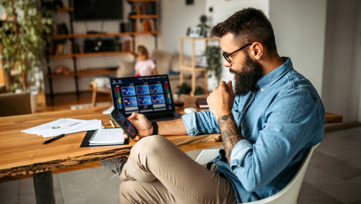 Ein Mann sitzt an einem Tisch auf dem ein aufgeklappter Laptop mit Aktienkursen auf dem Display steht. In der Hand hält er ein Smartphone und sieht sich ebenfalls kurse an.