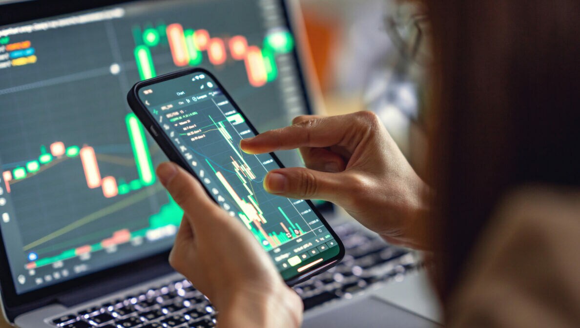 Nahaufnahme einer Person, die auf dem Smartphone Aktienkurse checkt. Im Hintergrund ist ein aufgeklappter Laptop auf dessen Bildschirm auch Aktienkurse zu sehen sind.