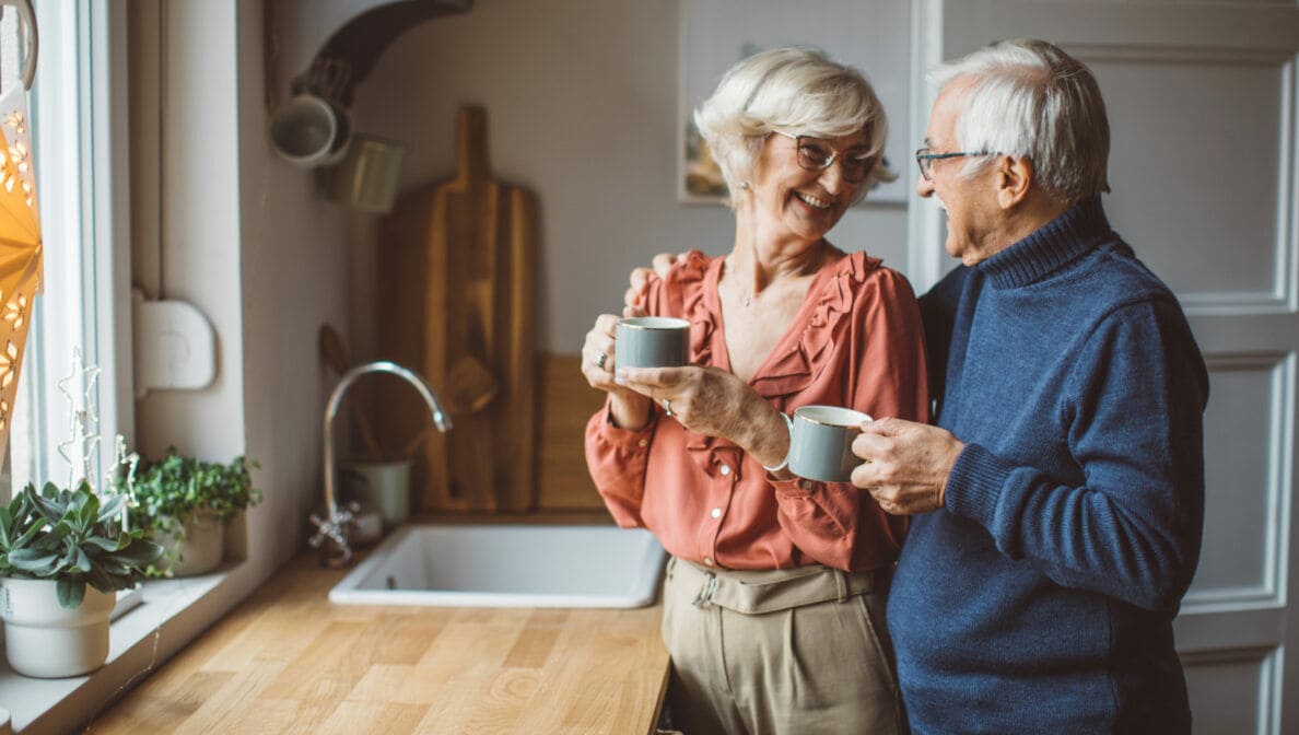 Zwei ältere Personen stehen in der Küche, halten Tassen in den Händen und blicken sich an.