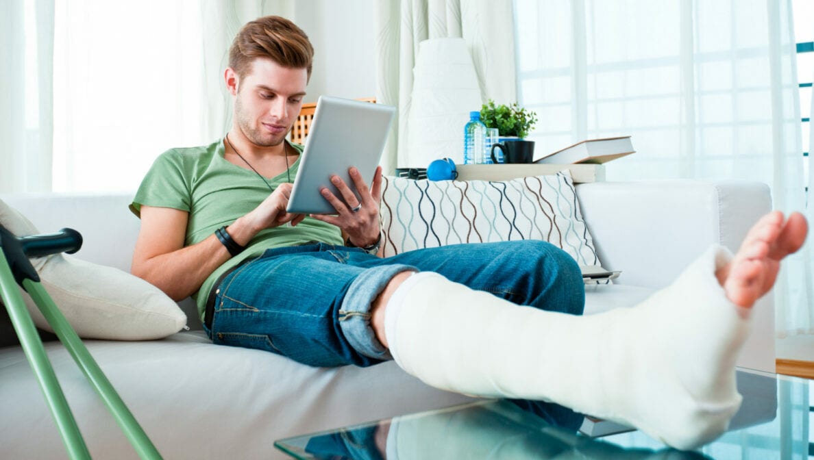 Eine Person mit gebrochenem Bein sitzt auf einem Sofa und schaut auf sein Tablet, neben ihr stehen Krücken.