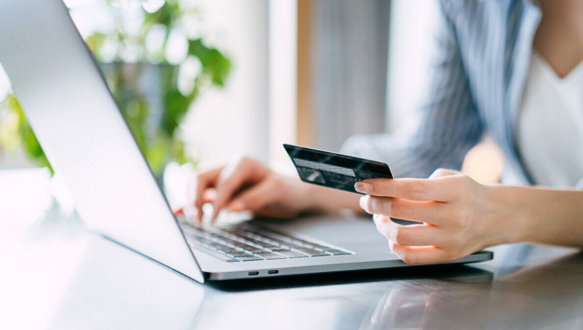 Nahaufnahme einer Hand, die eine Bankkarte hält, die andere Hand liegt auf einer Laptoptastatur.