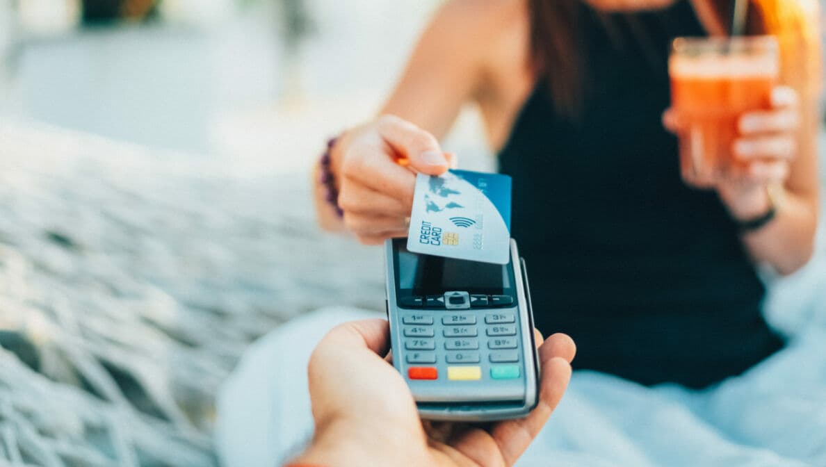 Eine Person in einer Hängematte hält ein Getränk in der Hand und bezahlt mit einer Kreditkarte.