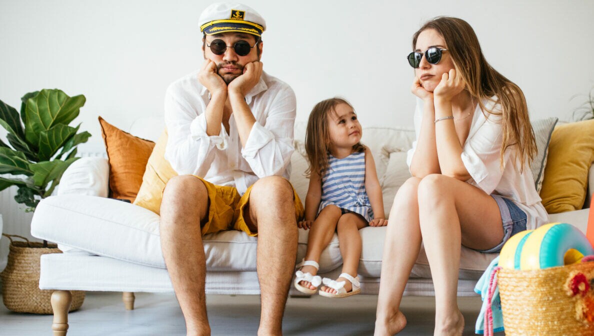 Eine Familie sitzt auf einem Sofa und stützt die Köpfe auf ihre Hände, daneben stehen gepackte Koffer.