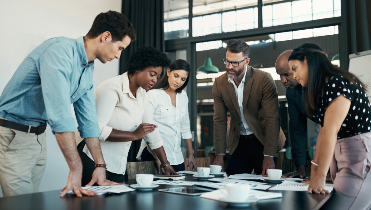 Sechs Personen stehen in einem Konferenzraum um einen Tisch herum und schauen auf Unterlagen in der Mitte des Tisches