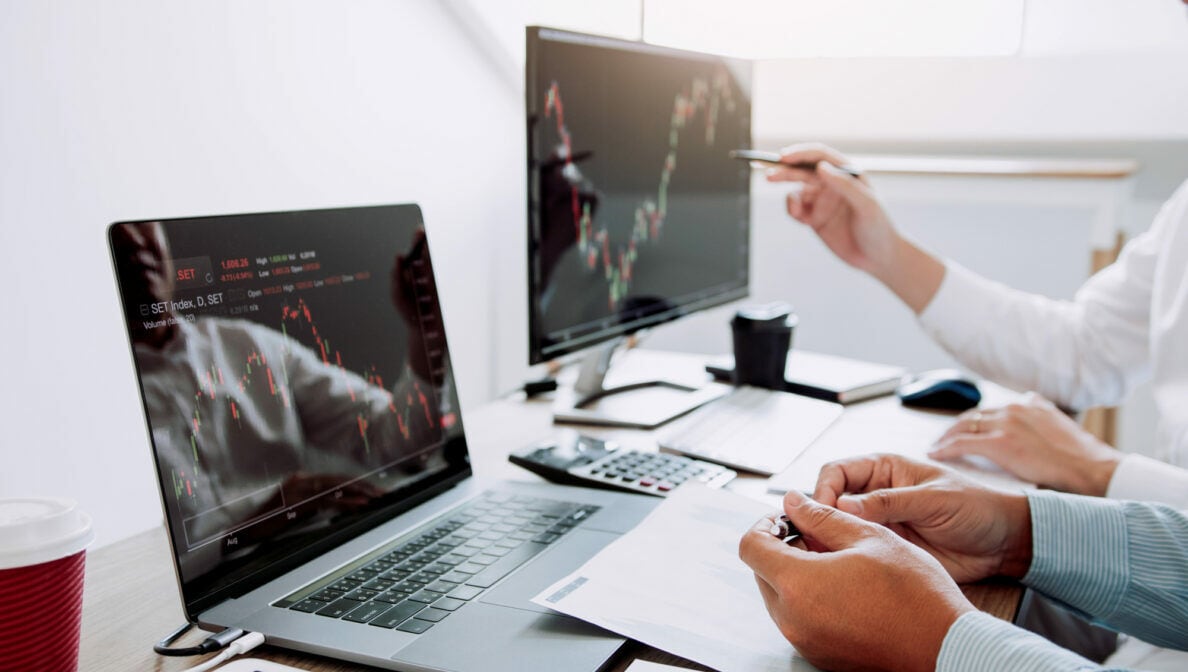 Zwei Personen sitzen vor einem Laptop und einem Monitor und betrachten Aktienkurse.