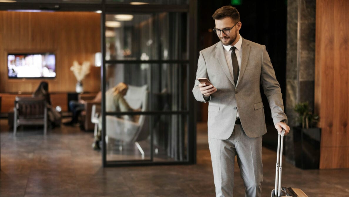 Eine Person im Businesslook und Trolley läuft mit Smartphone in der Hand durch eine Hotellobby.