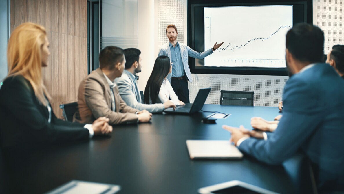 Mehrere Personen sitzen um einen Konferenztisch, am Kopfende steht eine Person und trägt etwas vor.