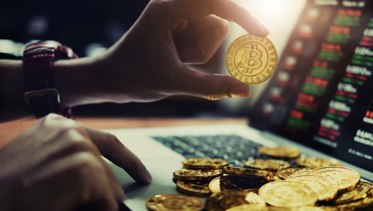 Ein Laptop mit Aktienkursen und einer Hand, die eine Bitcoin-Münze hält.