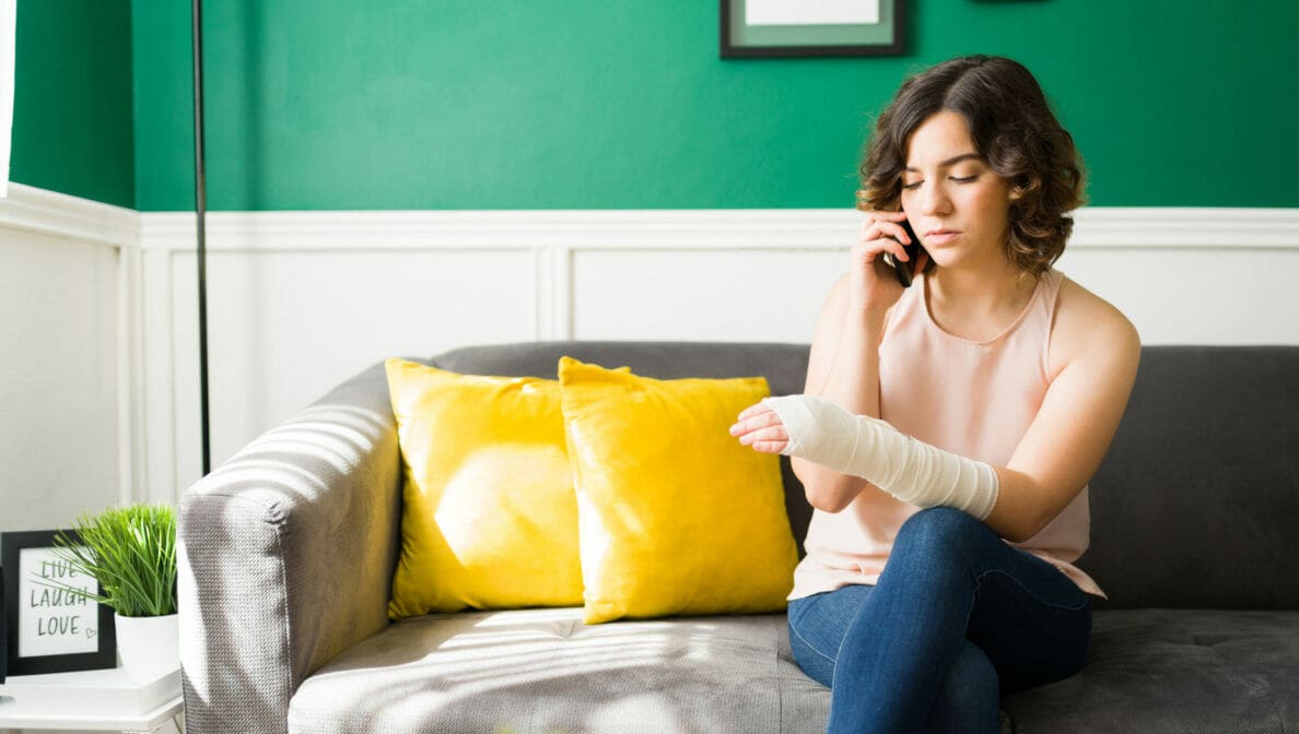 Eine Person sitzt mit einbandagiertem Arm auf einem Sofa und telefoniert.