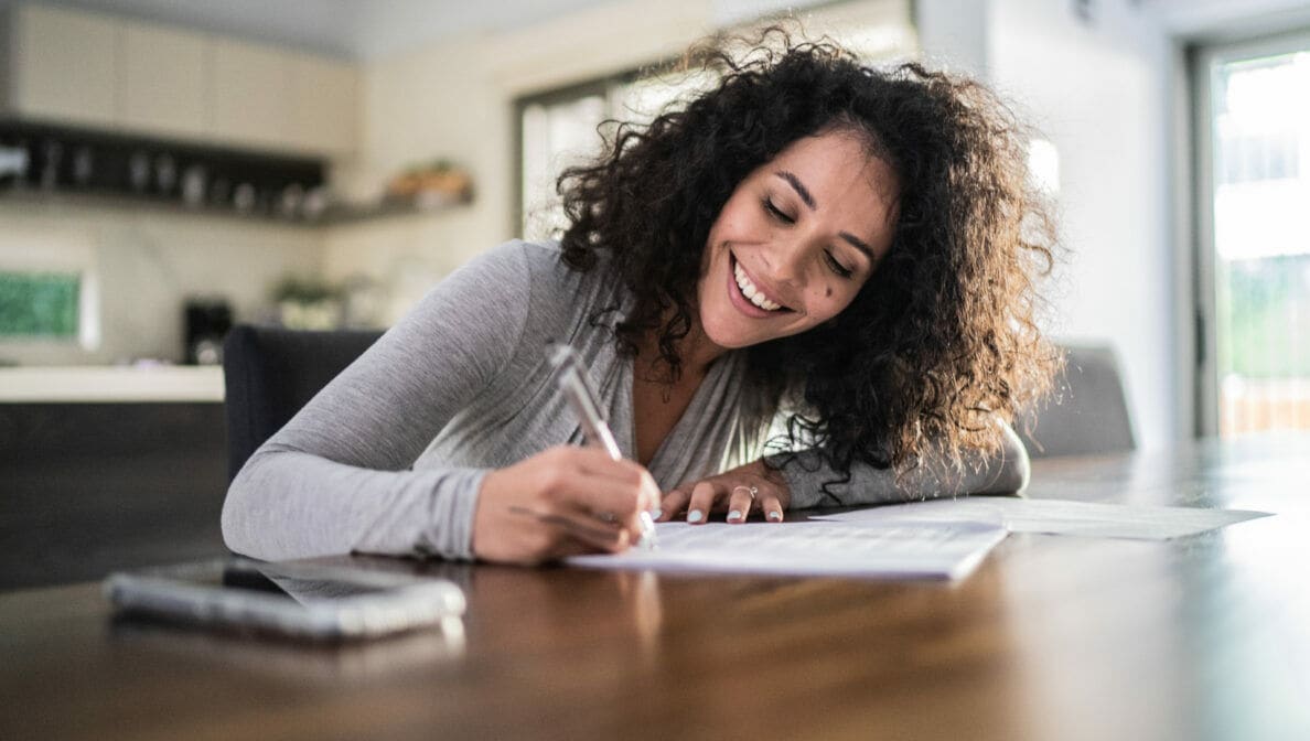 Eine Person sitzt am Tisch und trägt lächelnd etwas in ein Dokument ein.