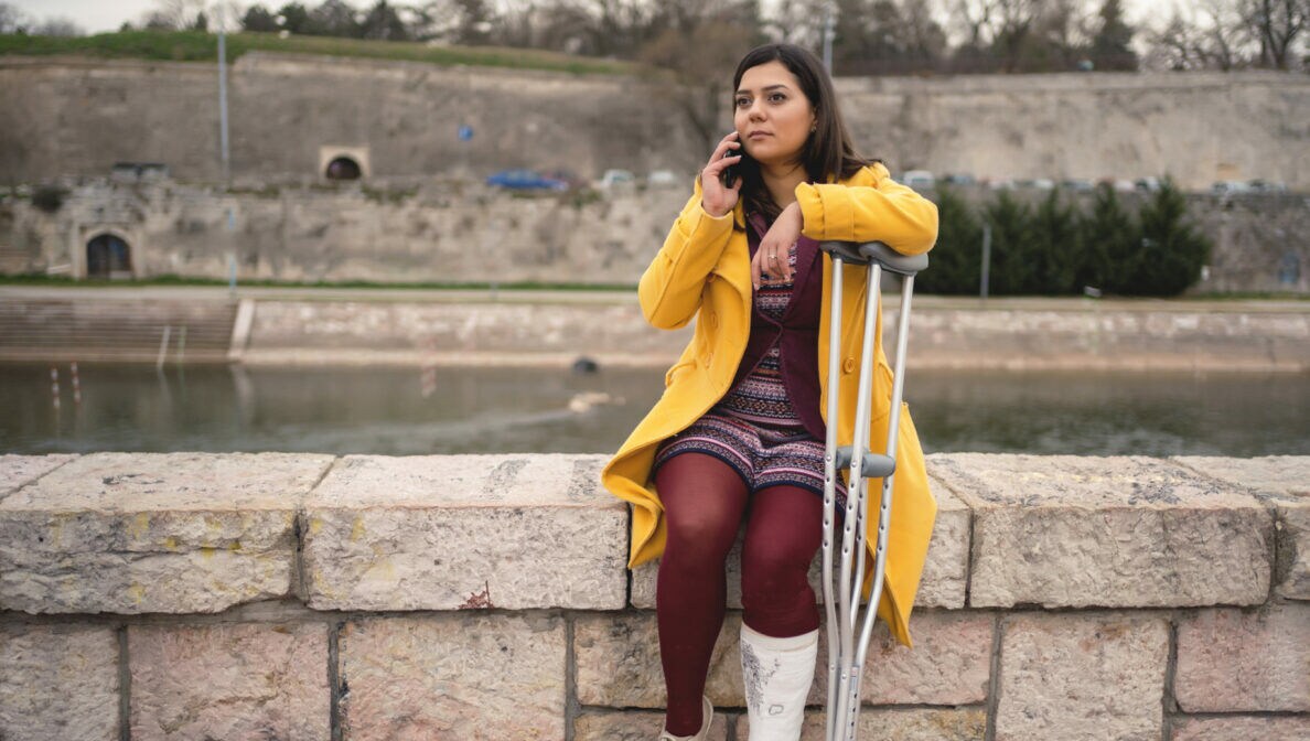 Eine Person mit eingegipstem Bein und Krücken sitzt auf einer Mauer am Fluss und telefoniert.