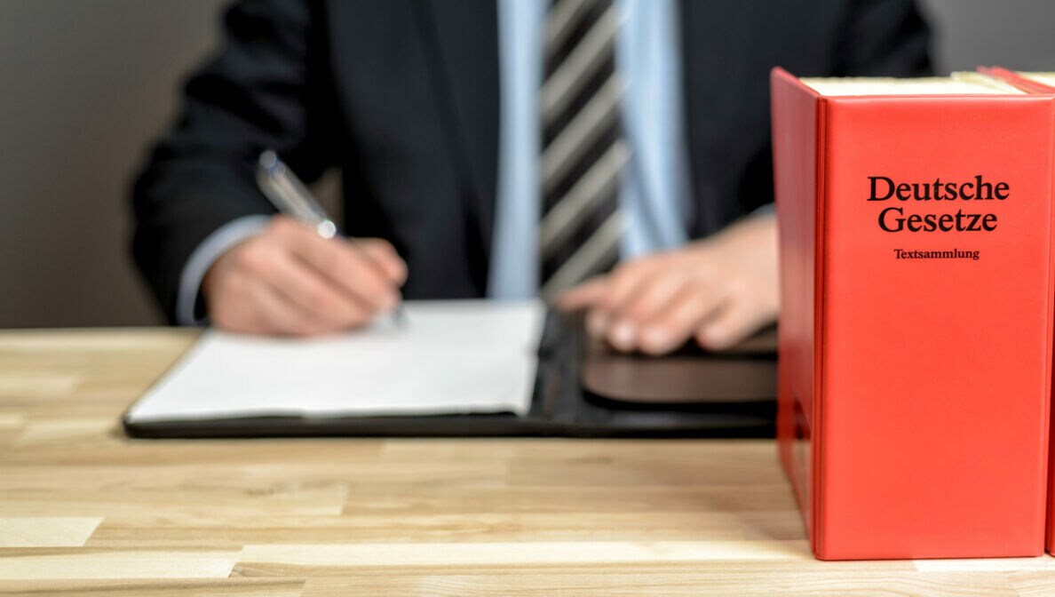 Eine Person im Anzug sitzt am Schreibtisch und unterzeichnet etwas, vor ihr stehen Gesetzesbücher.