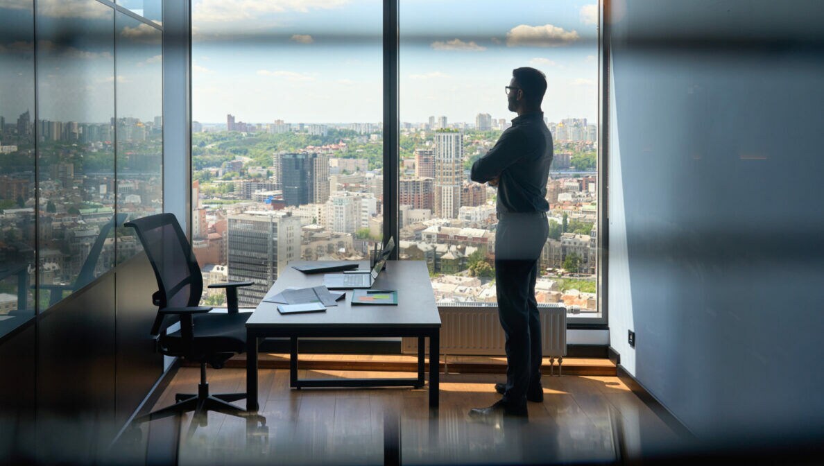Eine Person steht in einem Büro vor einer Fensterfront und blickt über das Stadtpanorama.