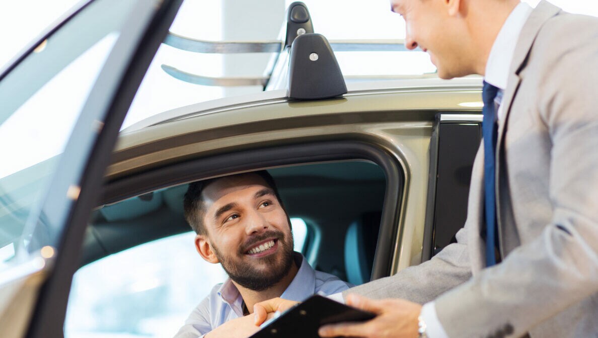 Eine Person sitzt im Autohaus in einem Auto und reicht einer anderen Person, die vor dem Auto steht, die Hand.