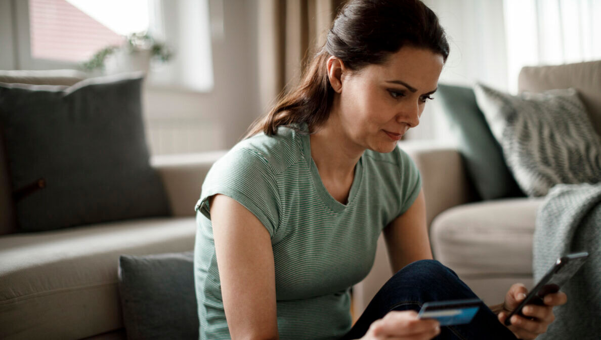 Eine Person sitzt auf dem Fußboden vor einem Sofa, hält in der einen Hand ein Smartphone und in der anderen Hand eine Bankkarte.