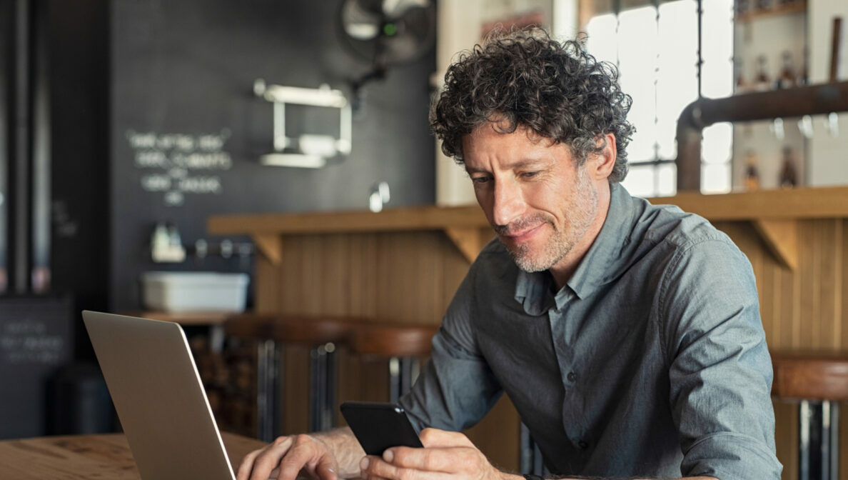 Eine Person blickt auf sein Smartphone und sitzt vor einem geöffneten Laptop.
