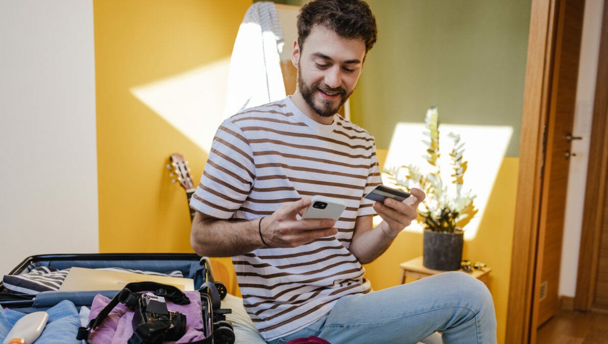 Eine Person sitzt neben einem aufgeklappten Koffer auf dem Bett und hält eine Bankkarte sowie ein Smartphone in den Händen.