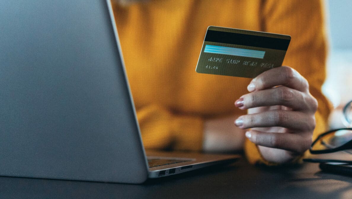 Nahaufnahme einer Person, die vor einem Laptop sitzt und eine Kreditkarte in der Hand hält.