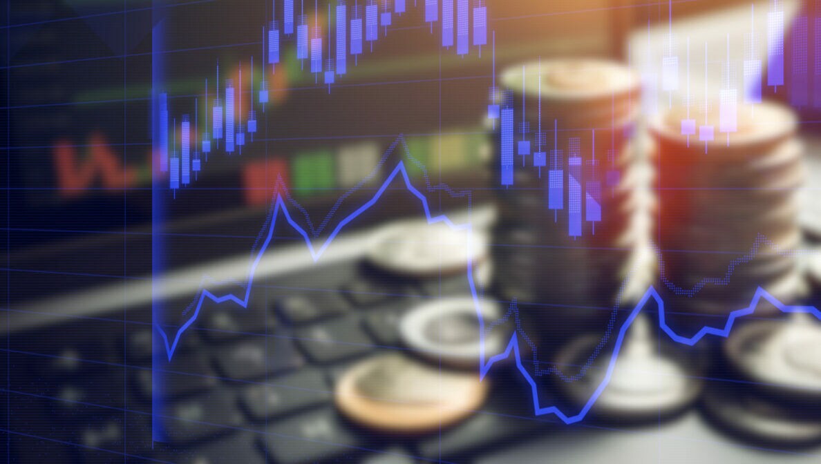 Im Vordergrund ist ein blau getönter Kerzenchart mit Indikatoren zu sehen. Im unscharfen Hintergrund sind auf einem Notebook Münzen aufgestapelt.