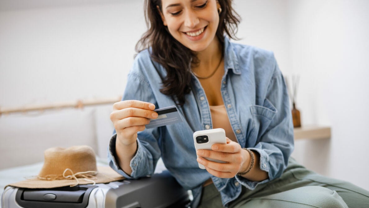 Eine Frau sitzt auf einen Koffer gelehnt und hält eine Kreditkarte und ein Smartphone in den Händen.