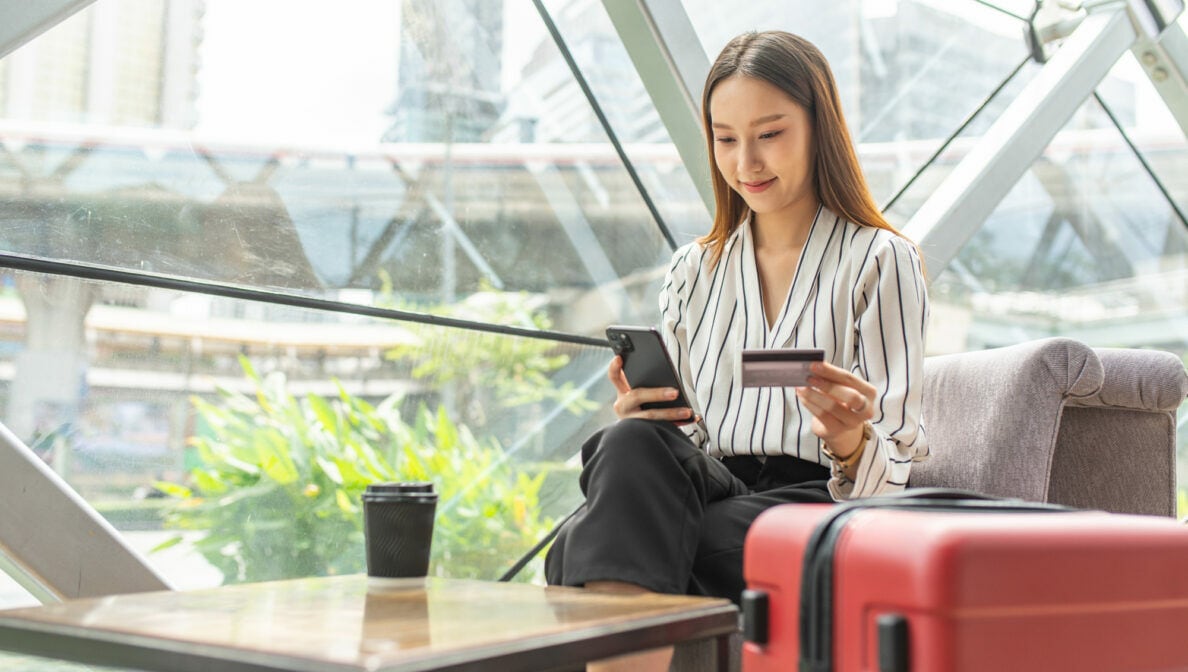 Eine Person sitzt mit einem Koffer neben sich an einem kleinen Beistelltisch und hält ein Smartphone sowie eine Bankkarte in den Händen.