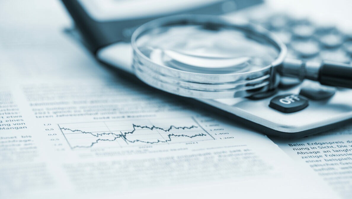 Nahaufnahme einer Lupe, die auf einem Taschenrechner und einem Papier mit Wirtschaftsinformationen liegt.