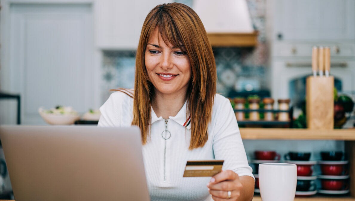 Eine Frau sitzt an einem Tisch vor einem aufgeklappten Laptop und hält eine Kreditkarte in der Hand.