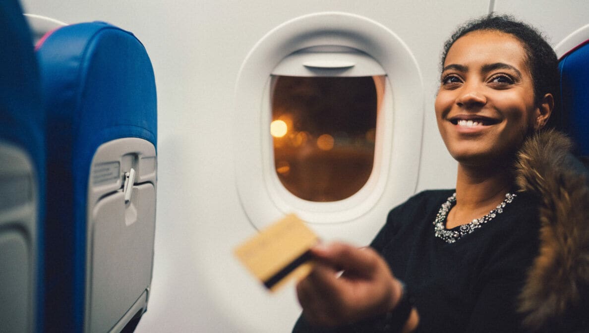 Eine Frau sitzt im Flugzeug und hält eine goldene Bezahlkarte hoch.
