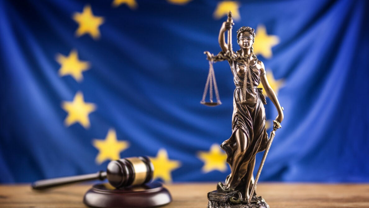 Eine Figur der Justitia steht auf einem Holztisch vor der europäischen Flagge, neben ihr liegt ein Richterhammer.