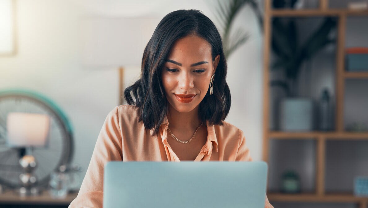 Eine lächelnde Frau sitzt an einem Tisch vor einem aufgeklappten Laptop.