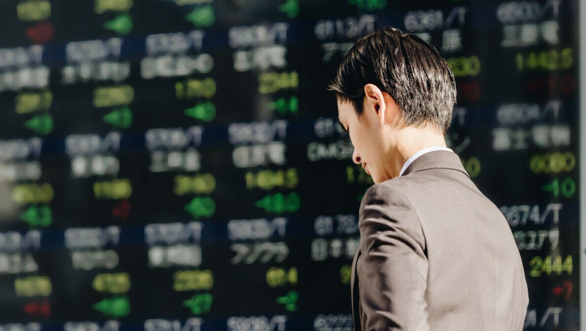 Eine Person im Anzug blickt auf ein Smartphone in ihrer Hand. Sie steht vor einer Anzeigetafel mit Wertpapierkursen.