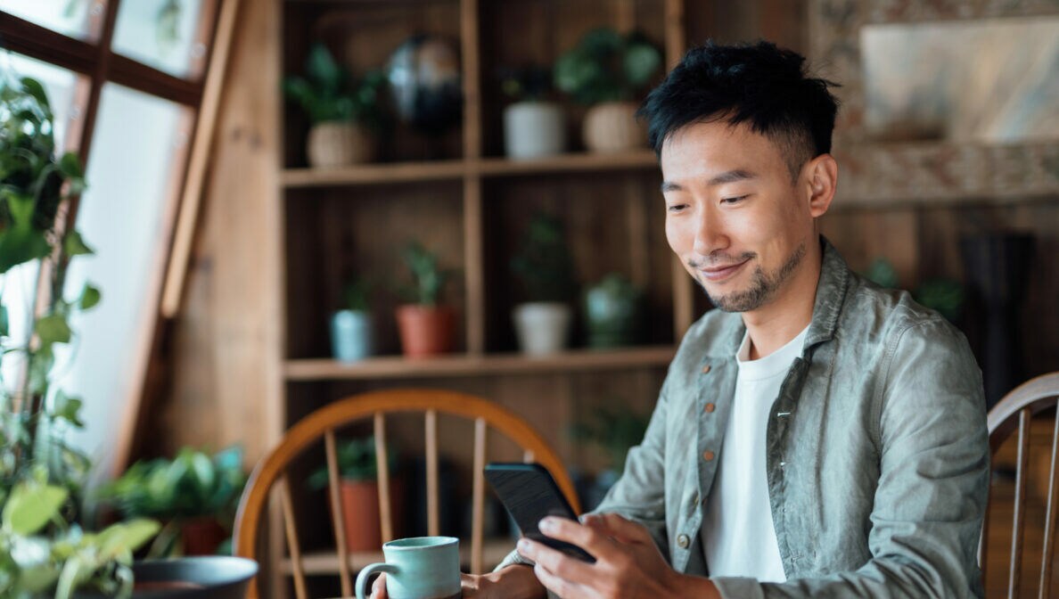 Eine Person sitzt an einem Tisch mit einer Kaffeetasse in der Hand und schaut auf ihr Smartphone in der anderen Hand.