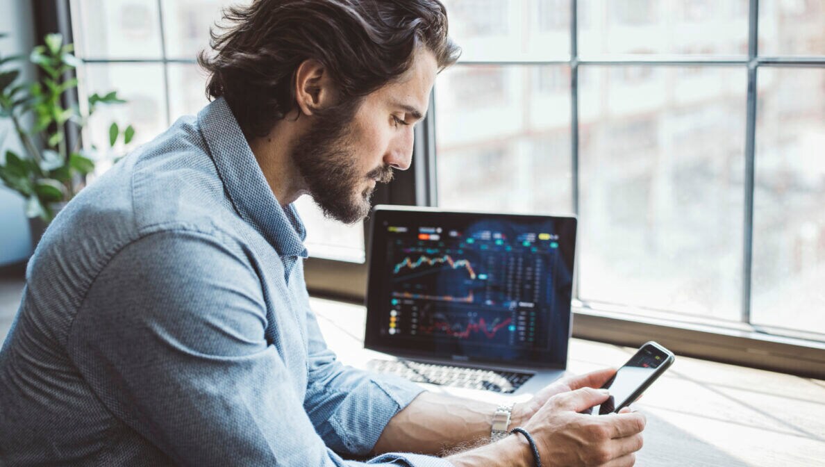 Ein Mann im hellblauen Hemd an einem Schreibtisch blickt auf das Smartphone in seiner Hand. Auf dem Tisch steht ein Notebook mit Börsenkursen auf dem Bildschirm.