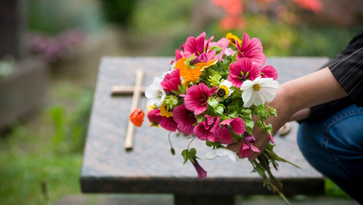 Eine Person legt einen Blumenstrauß vor einem Grabstein mit einem Kreuz nieder.