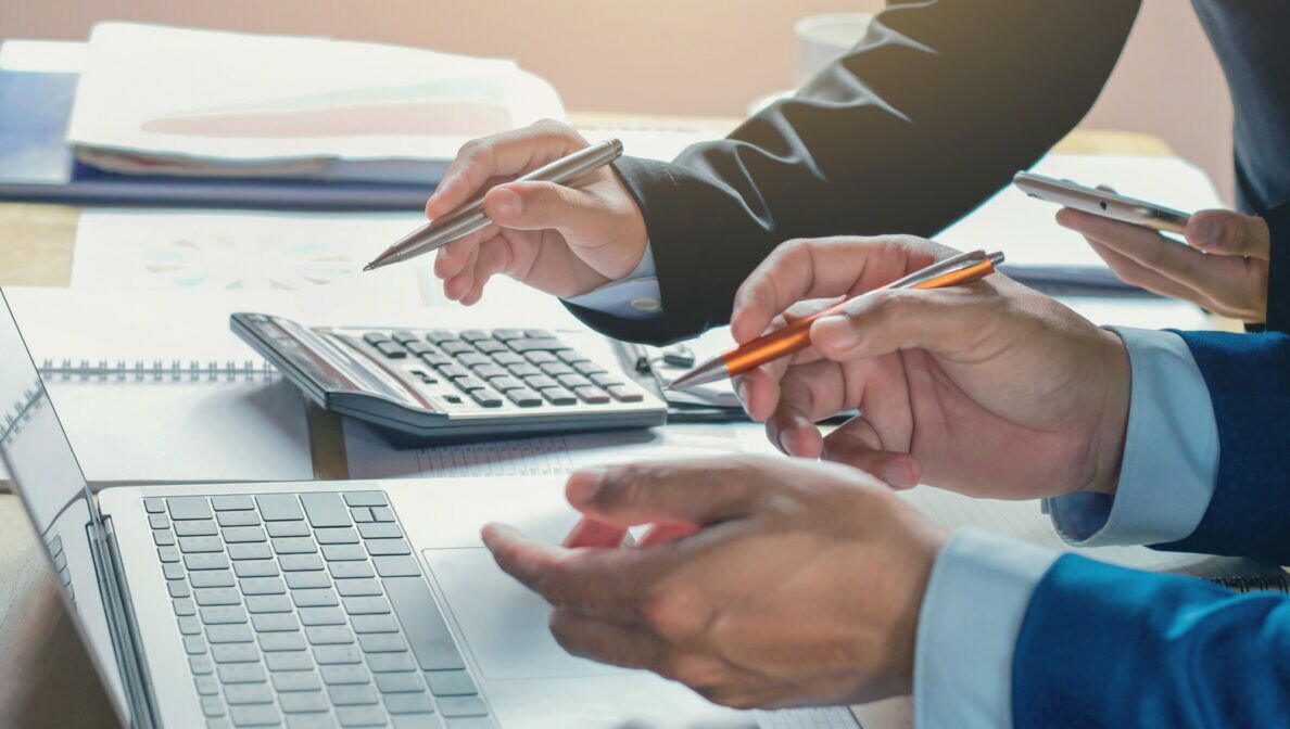 Zwei Personen mit Kugelschreiben in den Händen an einem Tisch mit geöffnetem Notebook, zahlreichen Papieren und einem Taschenrechner.