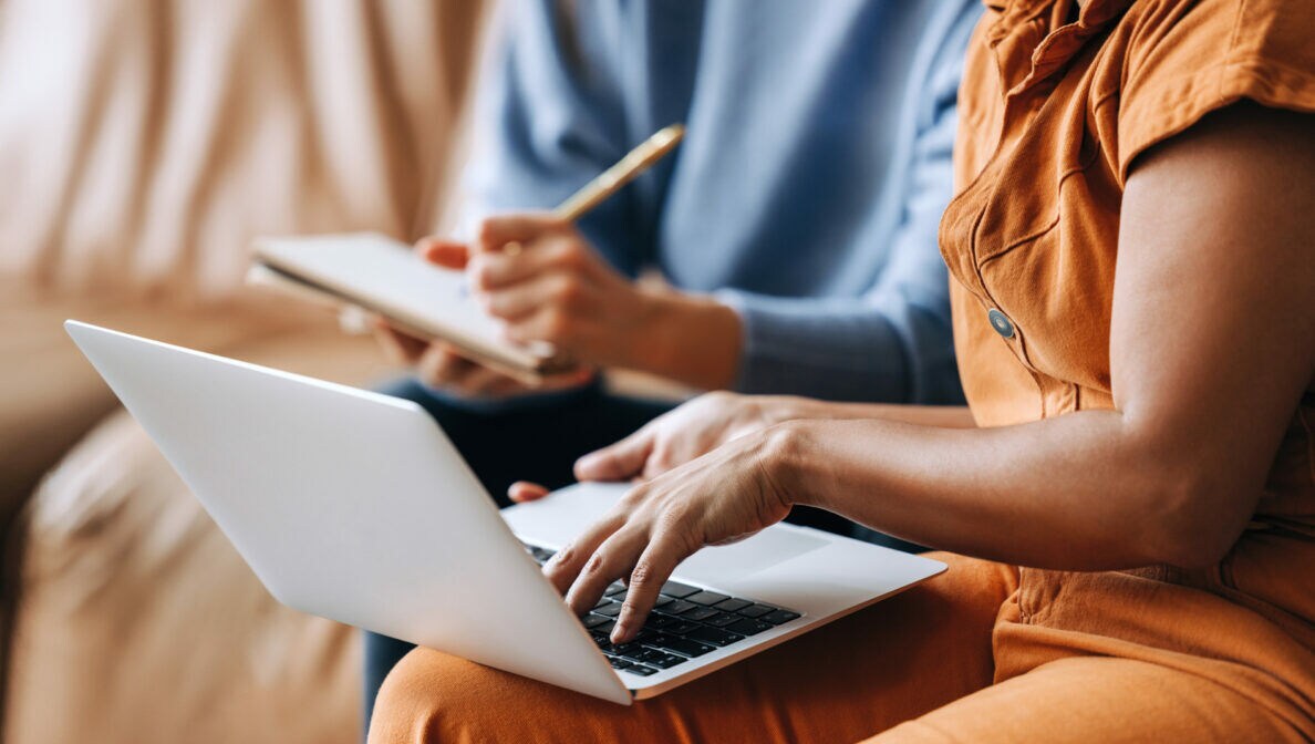 Eine Person hat einen Laptop auf dem Schoß und tippt auf der Tastatur, eine andere Person sitzt mit Notizblock und Stift daneben.