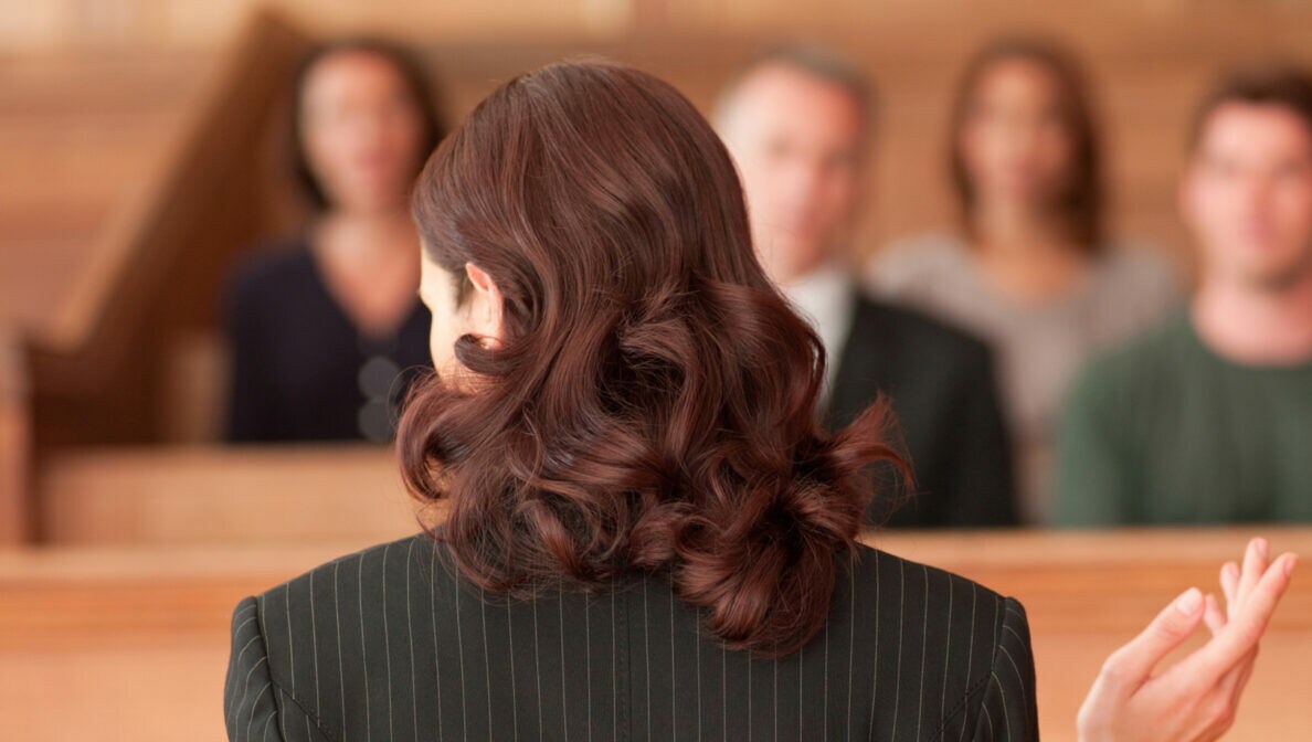 Eine Person im Gerichtssaal mit langen, rotbraunen Haaren in Nadelstreifenoberteil steht mit Dokumenten in einer Hand Personen gegenüber, die in den Gerichtssaalreihen sitzen.