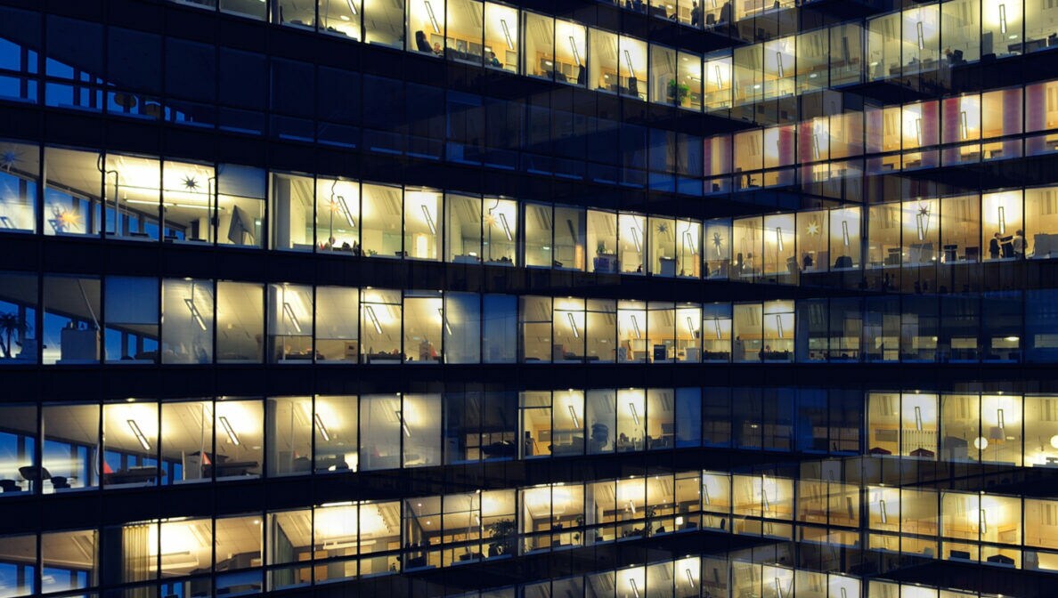 Außenansicht der Glasfassade eines beleuchteten Bürogebäudes im Abendlicht.