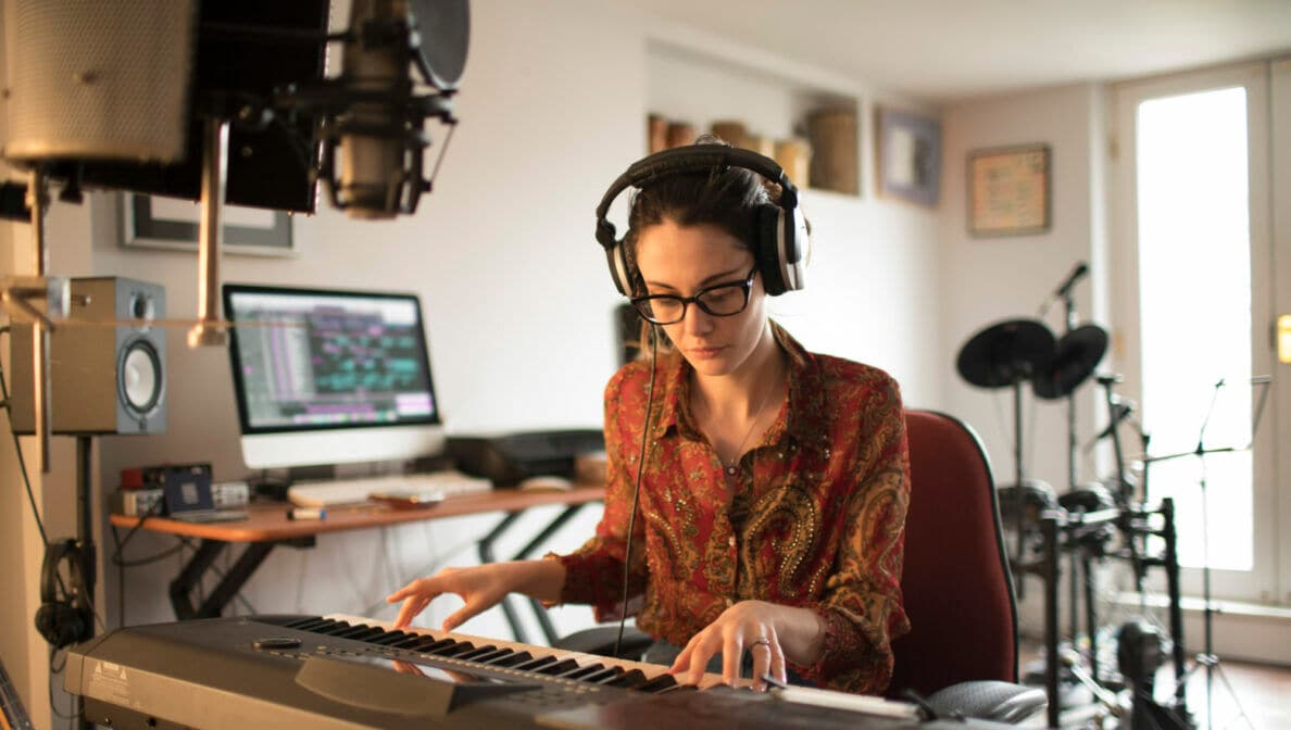 Eine Person sitzt in ihrer Wohnung an einem Keyboard, trägt Kopfhörer und ist von technischem Musikequipment umgeben.