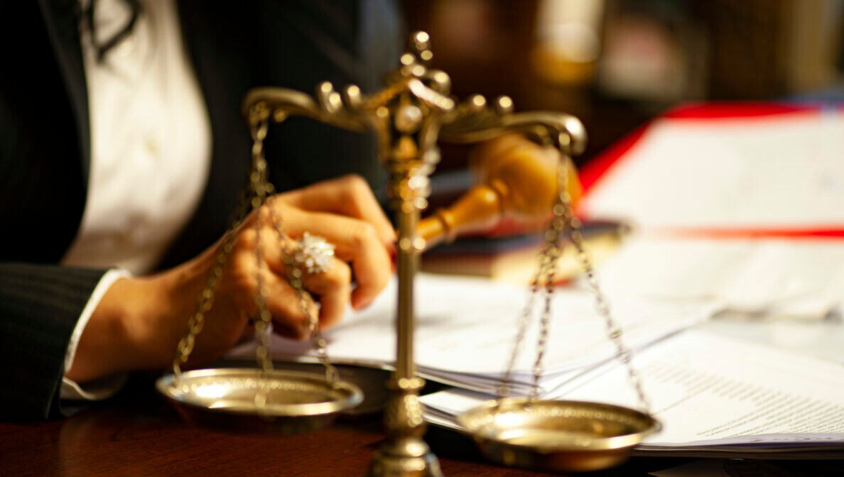 Eine Person an einem Tisch mit Goldwaage, Papieren und Ordnern hält einen Richterhammer in der Hand.