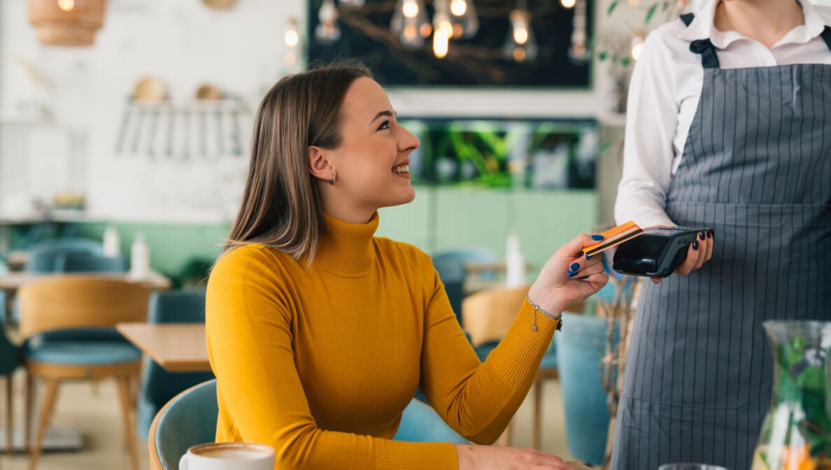 Eine Frau im gelben Rollkragenpullover an einem Tisch im Café mit einer Kaffeetasse hält eine Bezahlkarte an das Kartenlesegerät der Bedienung.
