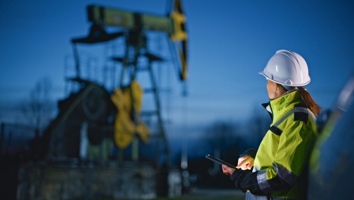 Eine Person in Berufskleidung mit Schutzhelm und Mobilgerät in der Hand schaut auf eine Ölförderanlage.