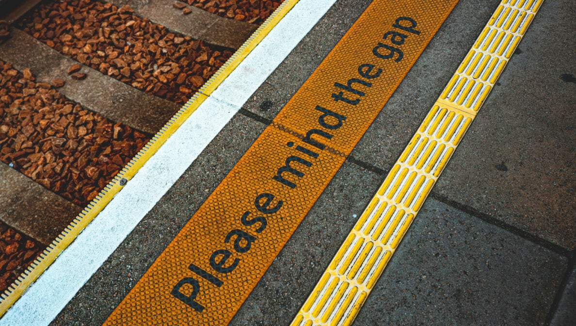 Der Sicherhinweis Please mind the gap auf dem Boden an einem Gleis.