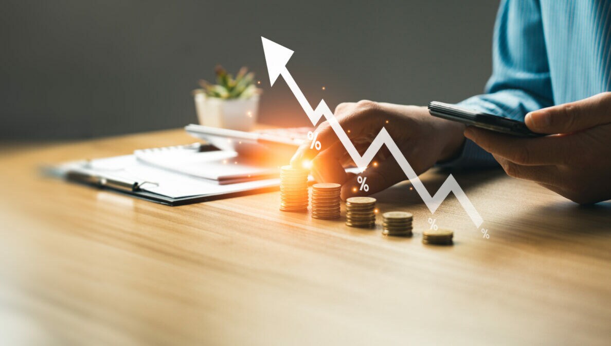 Eine Person an einem Tisch hält in einer Hand ein Smartphone, mit der anderen Hand stapelt sie Geld. Die Geldstapel sind mit einem nach oben steigenden Kursverlauf illustriert.