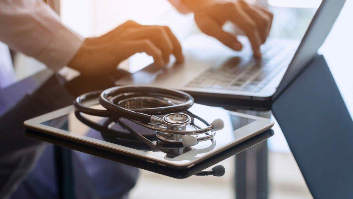 Eine Person tippt auf einem Laptop, daneben auf dem Glastisch liegt ein Stethoskop auf einem Tablet.