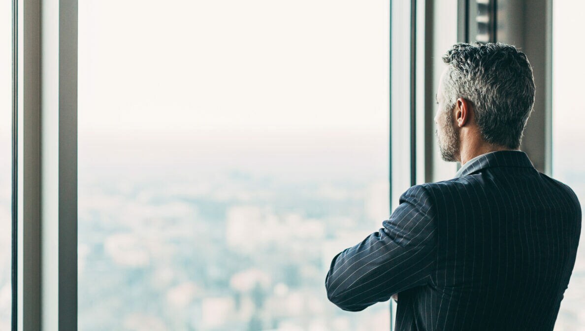 Eine Person im Anzug schaut mit verschränkten Armen aus einem großen Fenster in einer höher gelegenen Etage.