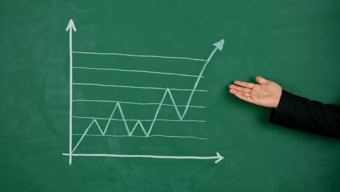 Eine Person deutet mit der Hand auf eine Grafik aus Kreide auf einer Tafel. Die Grafik zeigt ein Diagramm mit auf- und absteigendem Kurs, der am Ende steil nach oben deutet.