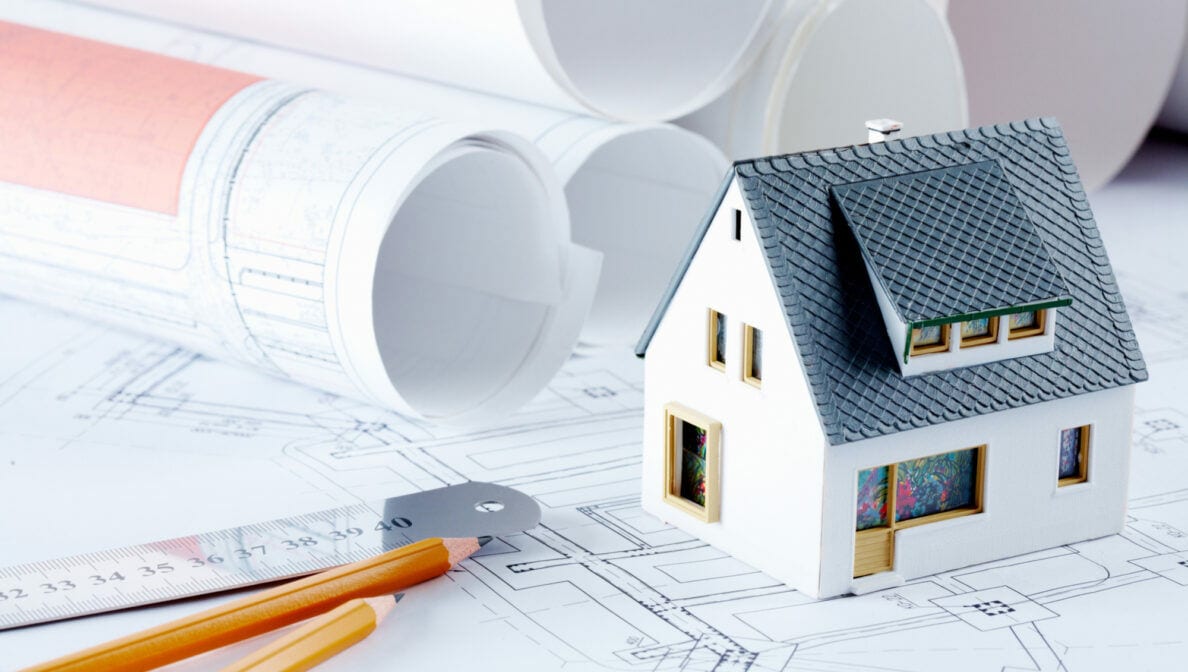 Ein Modell eines Hauses steht auf einem Grundriss, auf dem auch Bleistifte, ein Lineal und gerollte Baupläne liegen.
